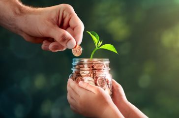 Hand Of Father Adding Penny To Child's Coin Jar With Plant Growing Out Of It - College Fund / Investing In Our Children's Future Concept