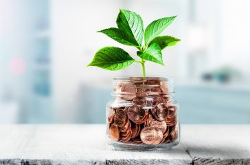 Plant Growing Out Of Coin Jar On Table In Office -  Investing / Business Success Concept