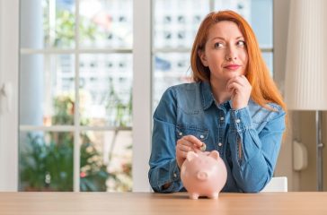 Redhead woman saves money in piggy bank at home serious face thinking about question, very confused idea