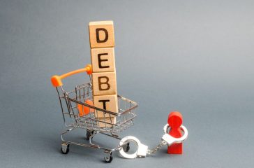Wooden blocks with the word Debt in a supermarket trolley and a human figurine bound by handcuffs. Mandatory payment of debt. Alimony payment. Pay of a mortgage loan. Debt load / obligation. IOU